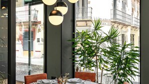 Une table de salle à manger ronde, recouverte d’une nappe blanche, agrémentée d’assiettes et de couverts, entourée de chaises en cuir, avec des lampes suspendues et des plantes en pot.