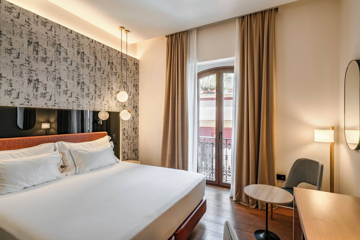 Habitación de hotel con una cama grande, un escritorio y un balcón con cortinas.
