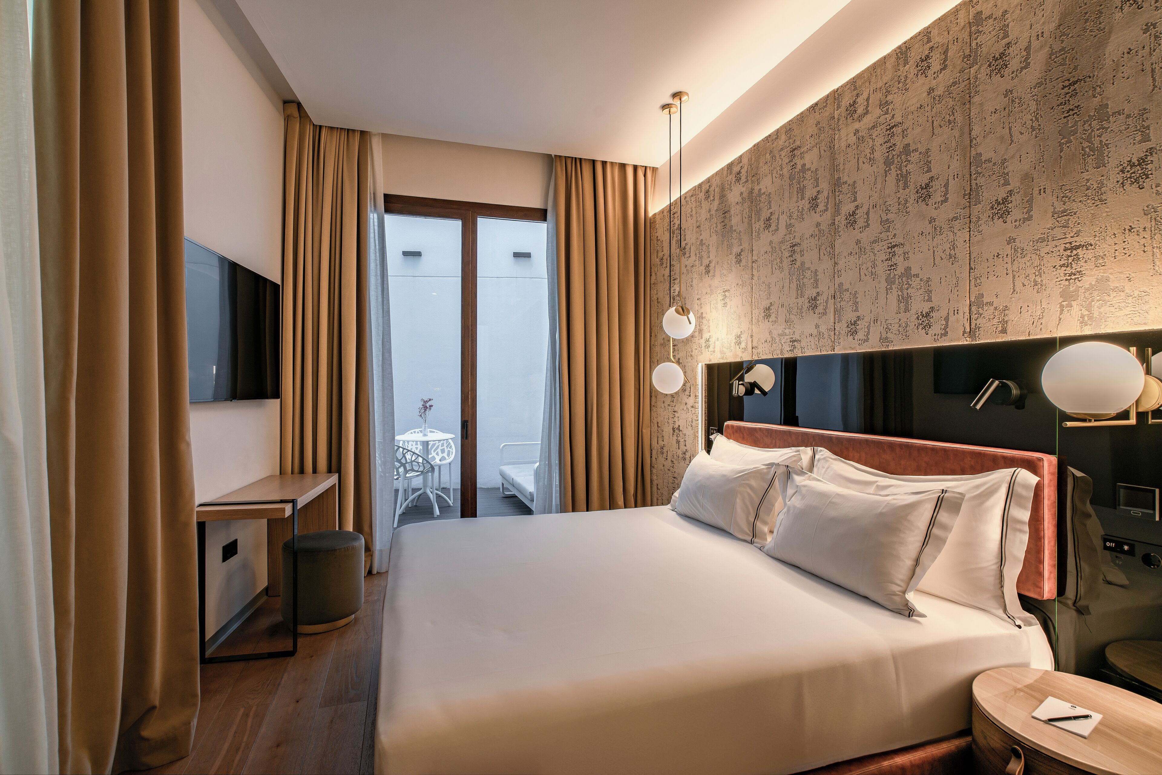A hotel room with a bed, bedside table, TV, and a small balcony with a table and chairs.