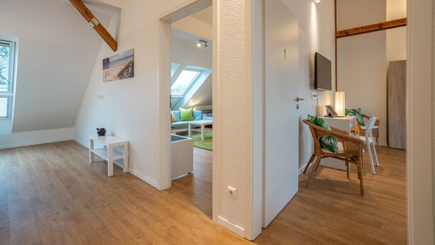 Un intérieur moderne avec un plancher en bois, une porte blanche, un coin repas avec des chaises en osier et un téléviseur bien visible.