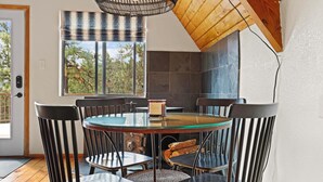 A dining area with a round glass table surrounded by six chairs, a wooden ceiling, and a window with a view of trees.