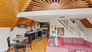 A cozy loft-style living space with a wooden ceiling, a dining area with a round table and chairs, a kitchenette, and a red plaid sofa.