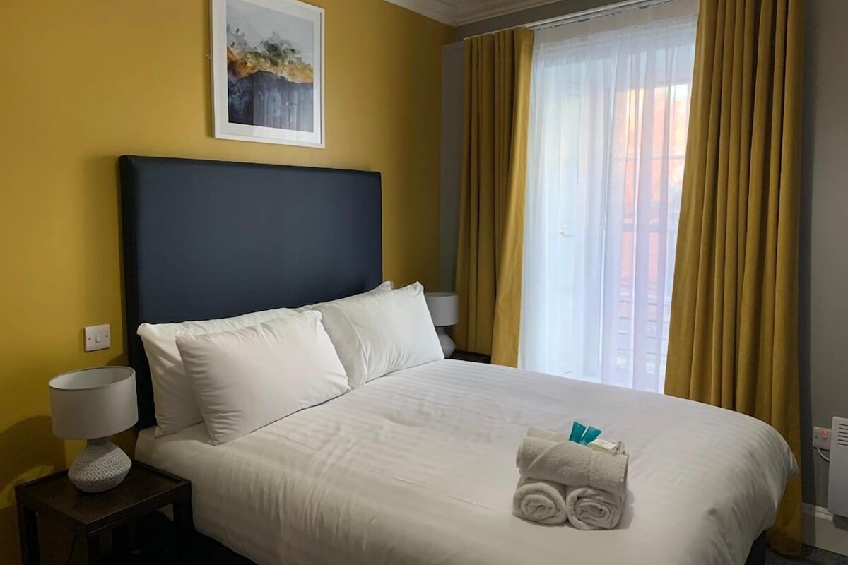 Habitación de hotel con una cama, una mesita de noche con lámpara, una ventana con cortinas y un cuadro en la pared.