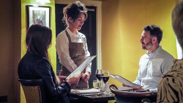 Interior de restaurante con una pared amarilla, un cuadro enmarcado y una mesa con copas de vino y una carta.