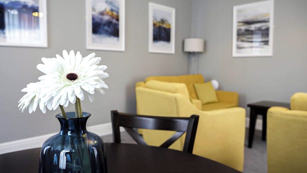 A room with a dining table, a vase with a white flower, a yellow sofa, and framed pictures on the wall. - Denvir's of Downpatrick (Downpatrick)
