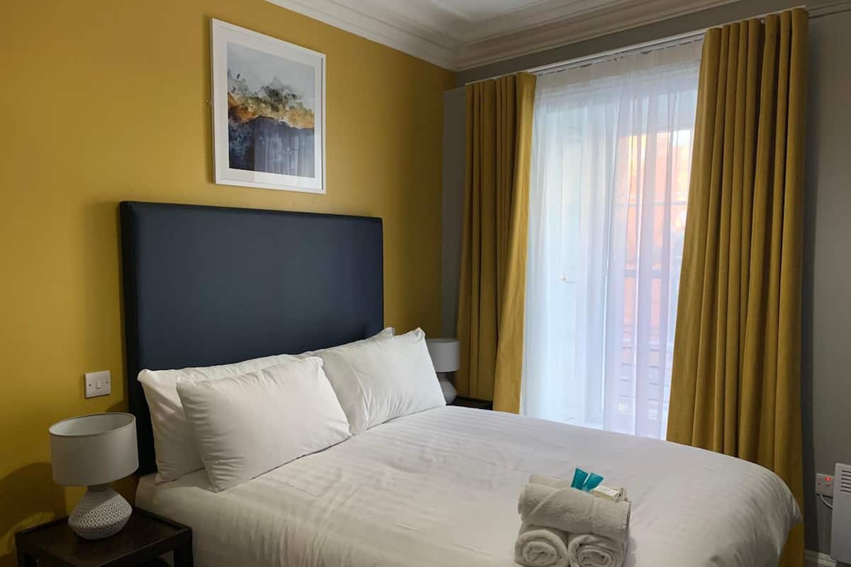 A hotel room with a large bed, a nightstand, a lamp, a framed picture on the wall, and a window with curtains.