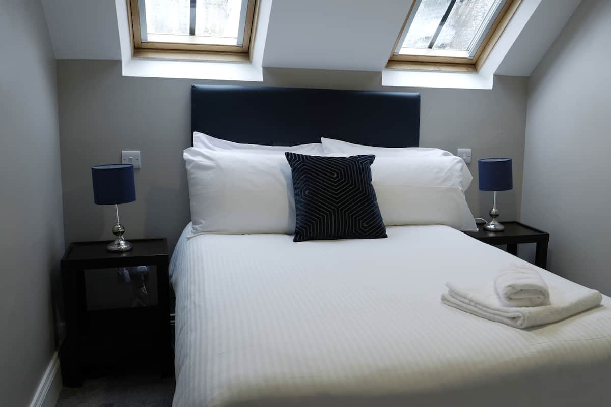 A neatly made bed with white linens and a folded towel, two bedside lamps, a dark headboard, and two skylights.