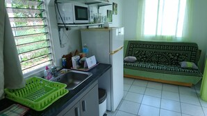 Kompakti keittiö, jossa on pesuallas, mikroaaltouuni ja jääkaappi. Työtasolla on vihreä kori ja taustalla kuvioitu päällinen sisältävä sohva.