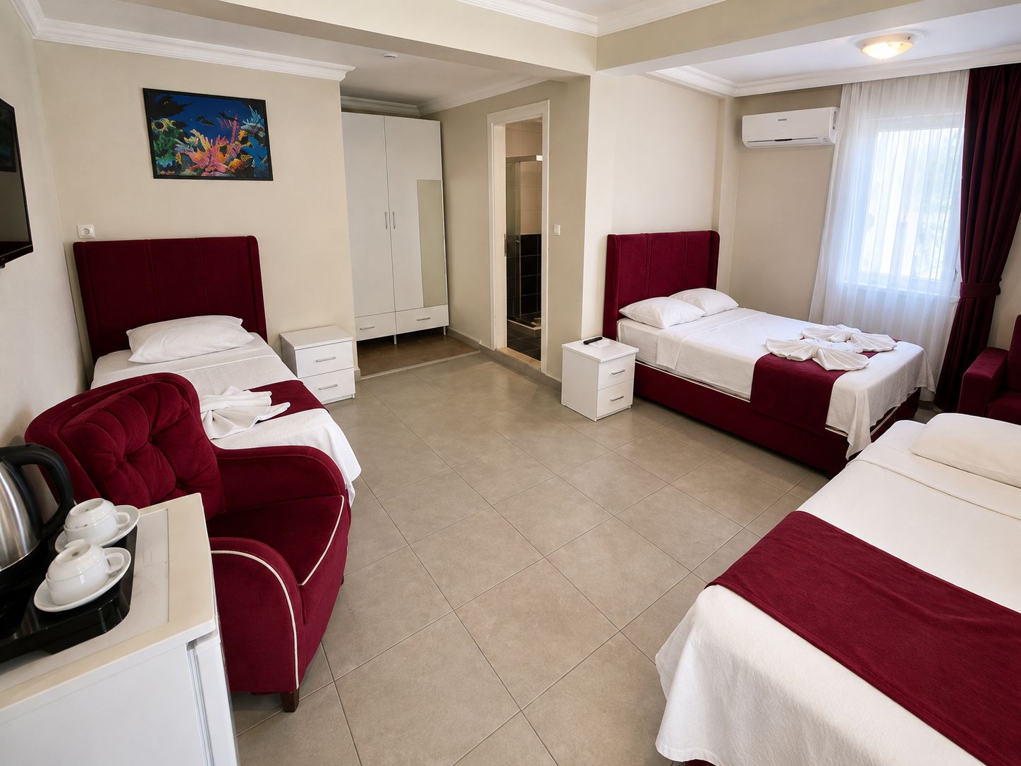 A hotel room with two beds, a seating area with a red sofa, a small table with a kettle and cups, and a painting on the wall.