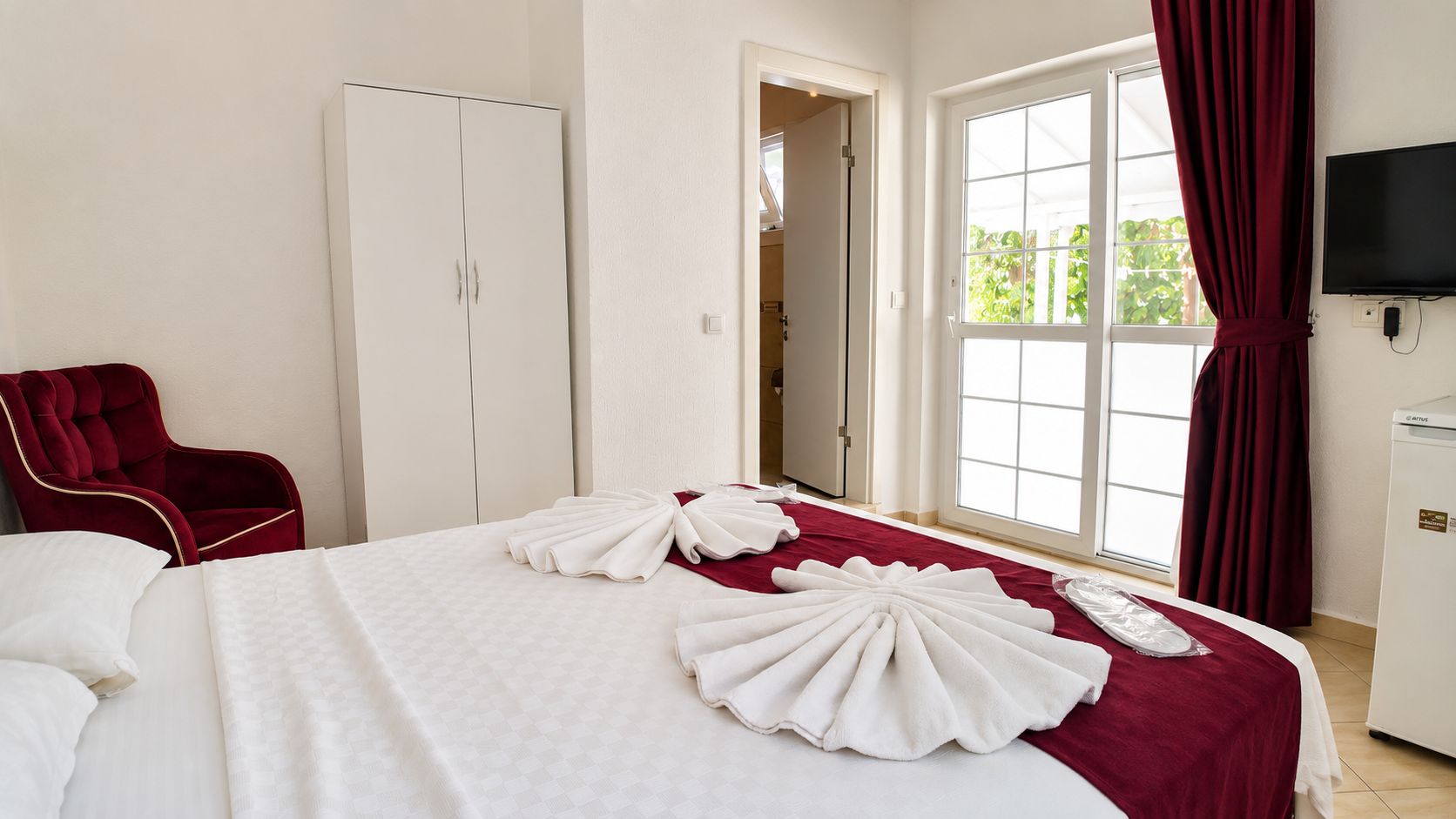 Una habitación de hotel con una cama, una silla roja, un armario, un televisor y un ventanal con cortinas.