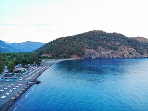 A coastal area with a beach lined with rows of sun loungers and umbrellas, a small building with a red roof, and a mountainous backdrop. - Bucak Apart Hotel Göcek - (Beach 1km)  (Fethiye)