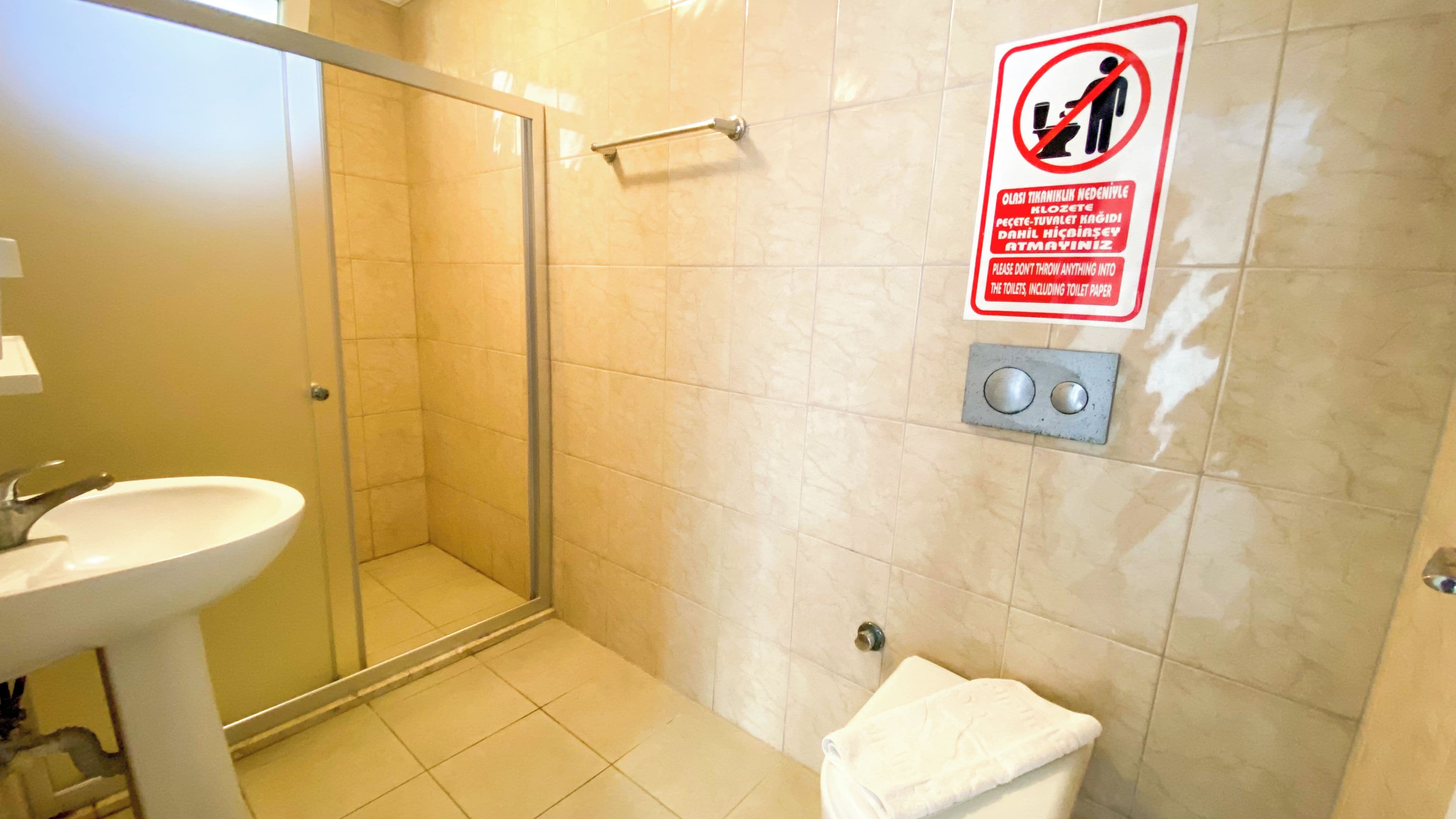 Un baño con puerta de ducha de vidrio, un lavamanos y un inodoro. Hay un letrero en la pared con un círculo rojo y una figura que no usa el inodoro.