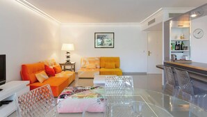 Una sala moderna con dos sofás naranja, una mesa de centro de vidrio, un televisor y una zona de bar con un reloj y estanterías.