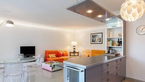 Una cocina moderna con una isla central, un comedor con sillas de vidrio transparente, un sofá con cojines naranja y un televisor de pantalla plana.