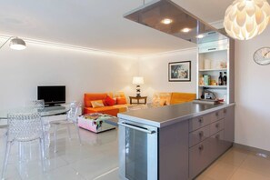 Una cocina moderna con una isla central, un comedor con sillas de vidrio transparente, un sofá con cojines naranja y un televisor de pantalla plana.
