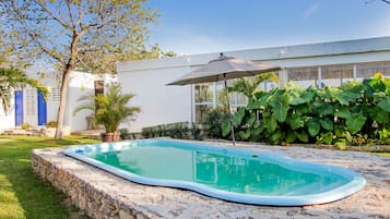 A swimming pool with a stone border, surrounded by a grassy area and a house with large windows and a covered patio.