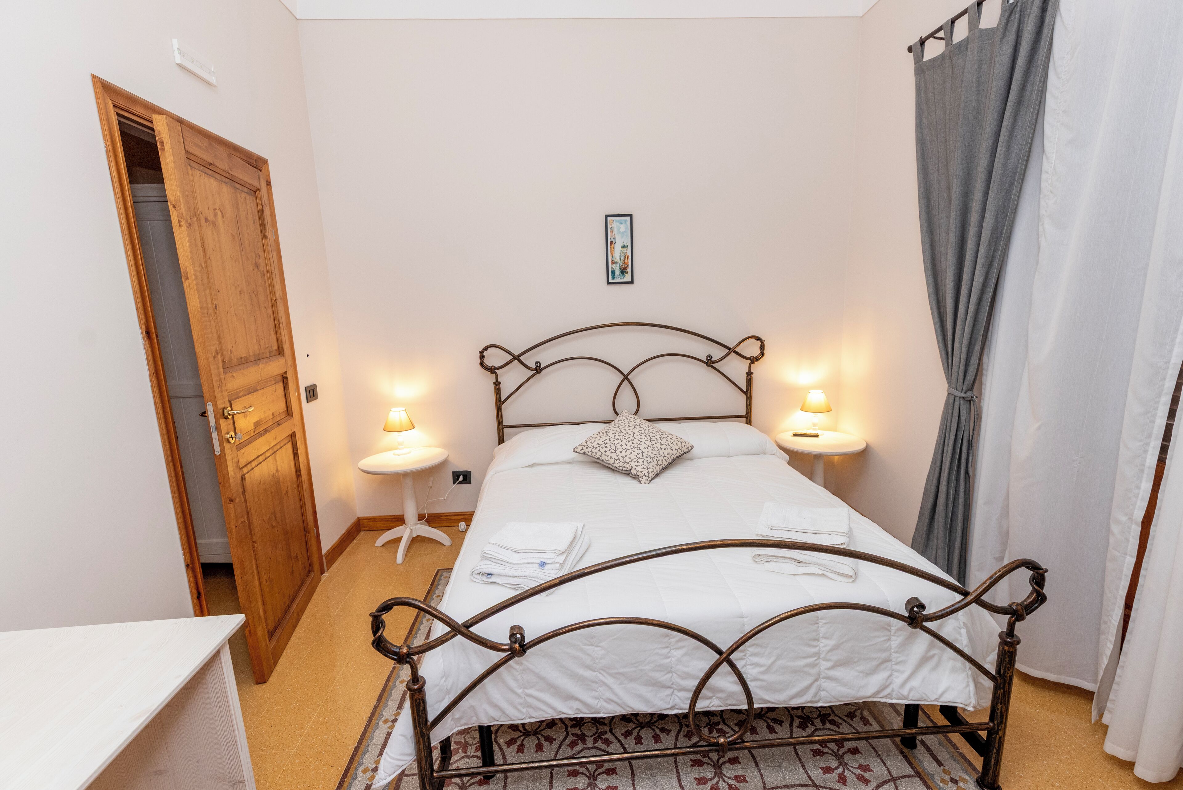 A neatly arranged bedroom with a metal bed frame, white bedding, two bedside lamps, a small bedside table, and a door leading to another room.