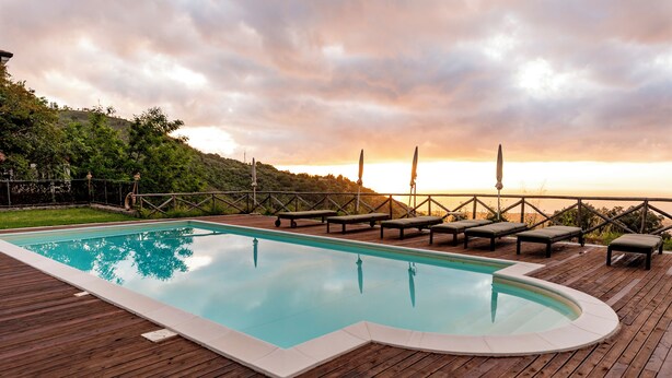 Um deck de madeira com piscina, cadeiras de descanso e vista para o pôr do sol sobre uma paisagem montanhosa.