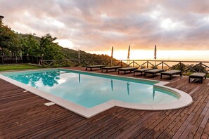 Una terraza de madera con piscina, sillas de descanso y vistas a un paisaje montañoso al atardecer.