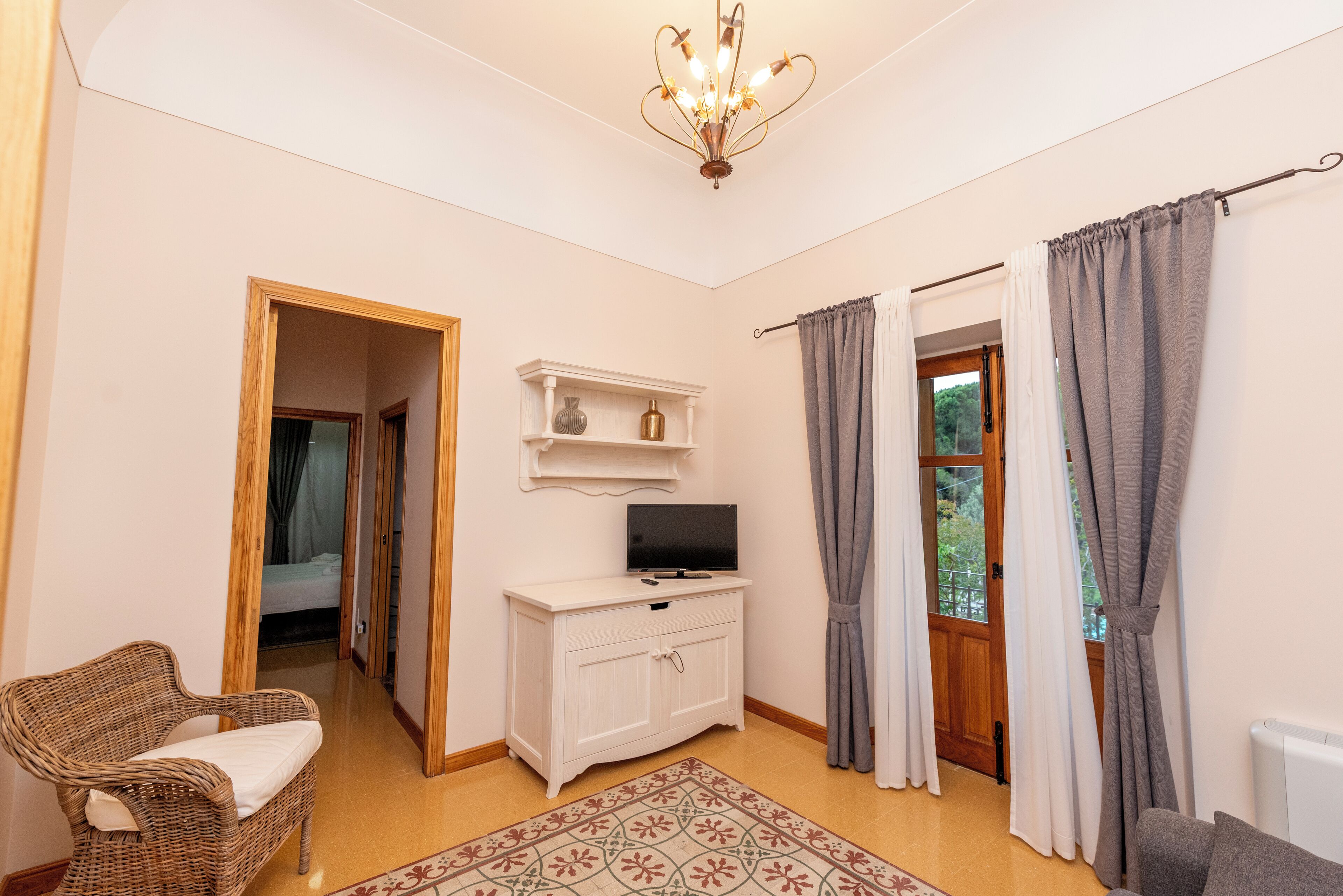 A room with a wicker chair, a white cabinet with a television, a patterned rug, and a door leading to a balcony.