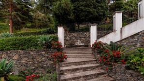 Uma escadaria de pedra leva a um edifício branco, cercado por vegetação exuberante e flores vermelhas.