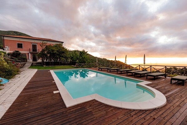 Terraza de madera con piscina, sillas de descanso y vista al atardecer sobre una colina.