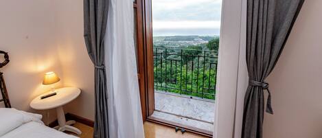Une chambre d’hôtel avec un lit, une petite table avec une lampe et une vue sur un paysage verdoyant, visible par la porte d’un balcon.