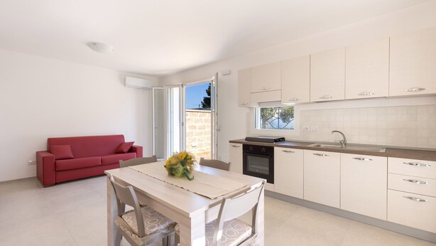 Een compacte leefruimte met een kitchenette, een eethoek met een tafel en stoelen, een rode bank en uitzicht door een geopende deur.