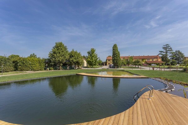 Une piscine dotée d’une terrasse en bois et de rampes en métal, entourée de verdure et avec des bâtiments en arrière-plan.