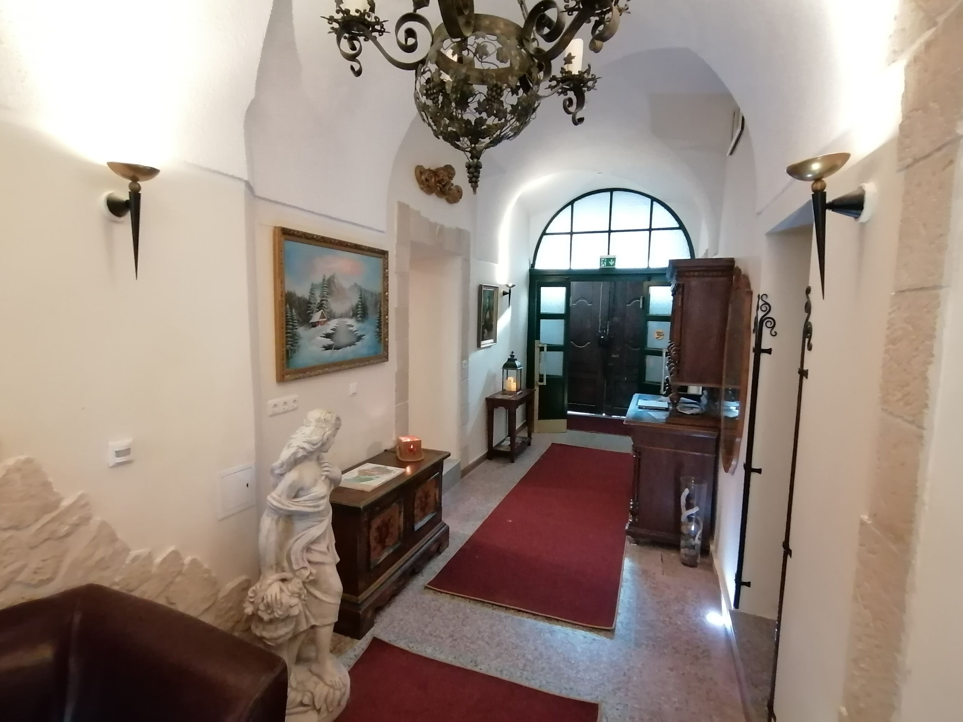 A hallway with a red carpet, a statue, a painting, a chandelier, and a wooden cabinet.