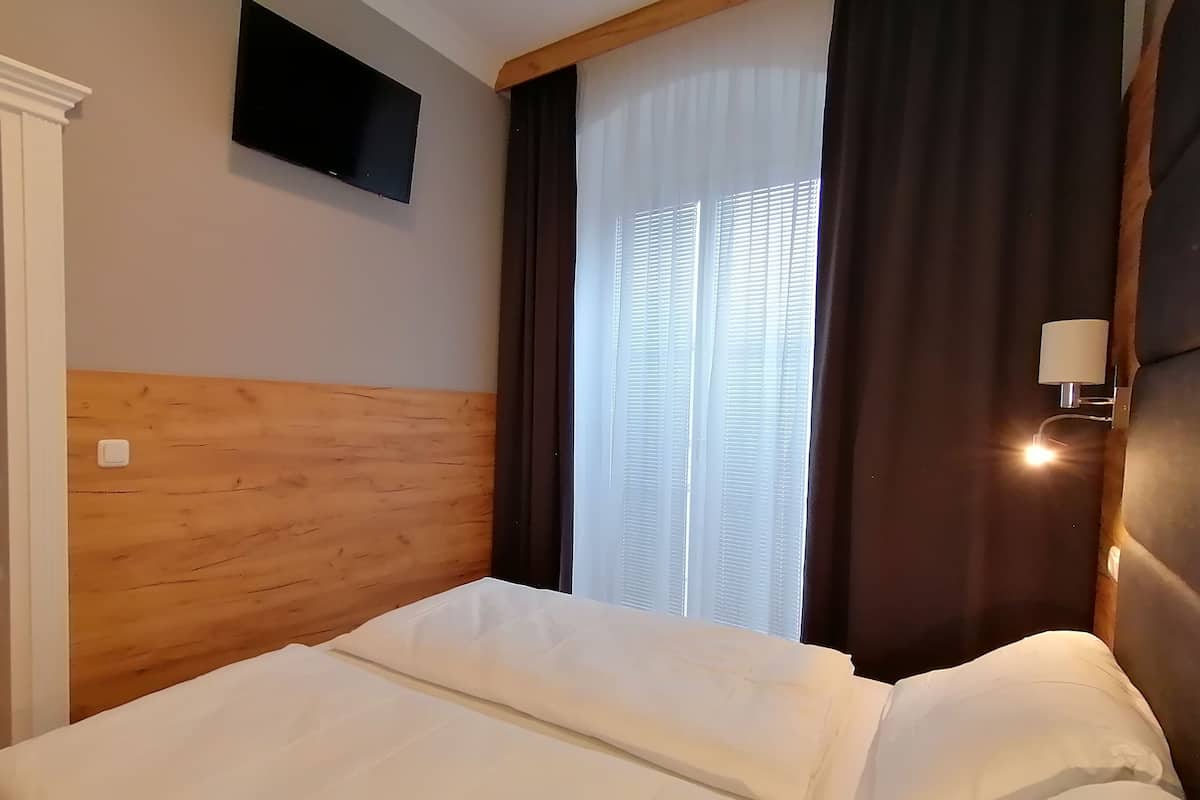 A hotel room with a wooden headboard, a flat-screen TV mounted on the wall, a window with blinds, and a bedside lamp.
