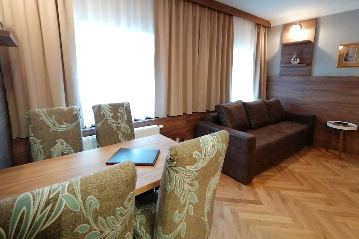 A hotel room with a wooden floor, a brown sofa, a dining table with four chairs, and a large window with curtains.