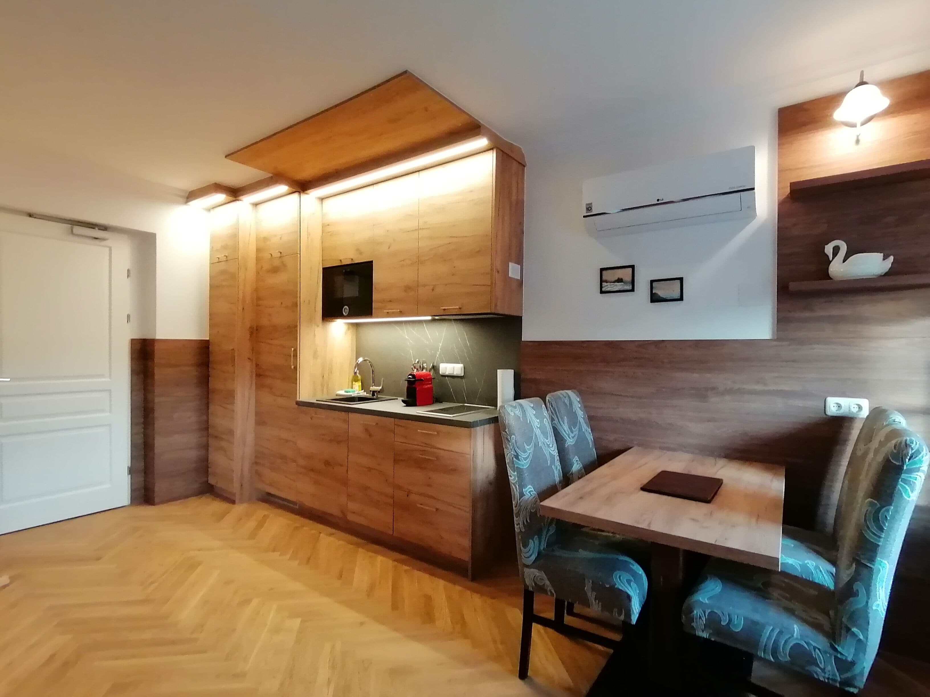 A compact kitchen with wooden cabinets, a built-in microwave, and a sink. There is a dining area with a table and chairs, and a wall-mounted air conditioning unit.