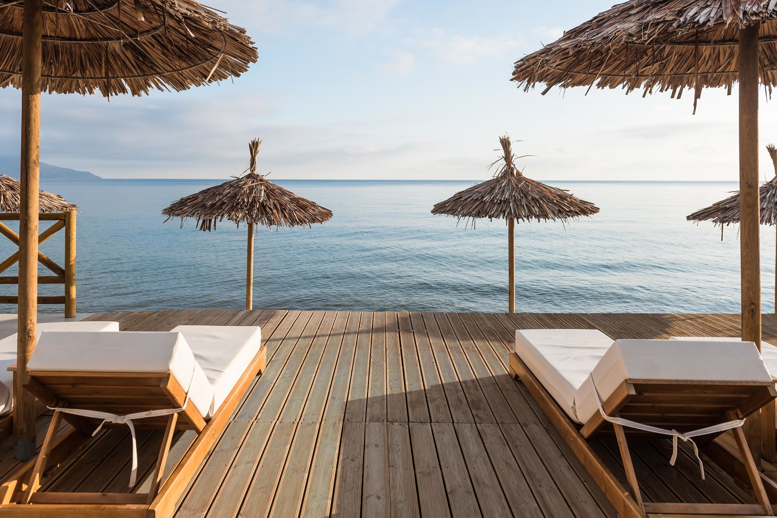 Una terraza de madera con sillones de descanso y sombrillas de paja que da a un mar tranquilo.