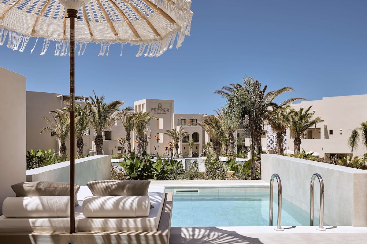 A pool area with a lounge chair and umbrella, surrounded by palm trees and a building labeled "PEPPER."