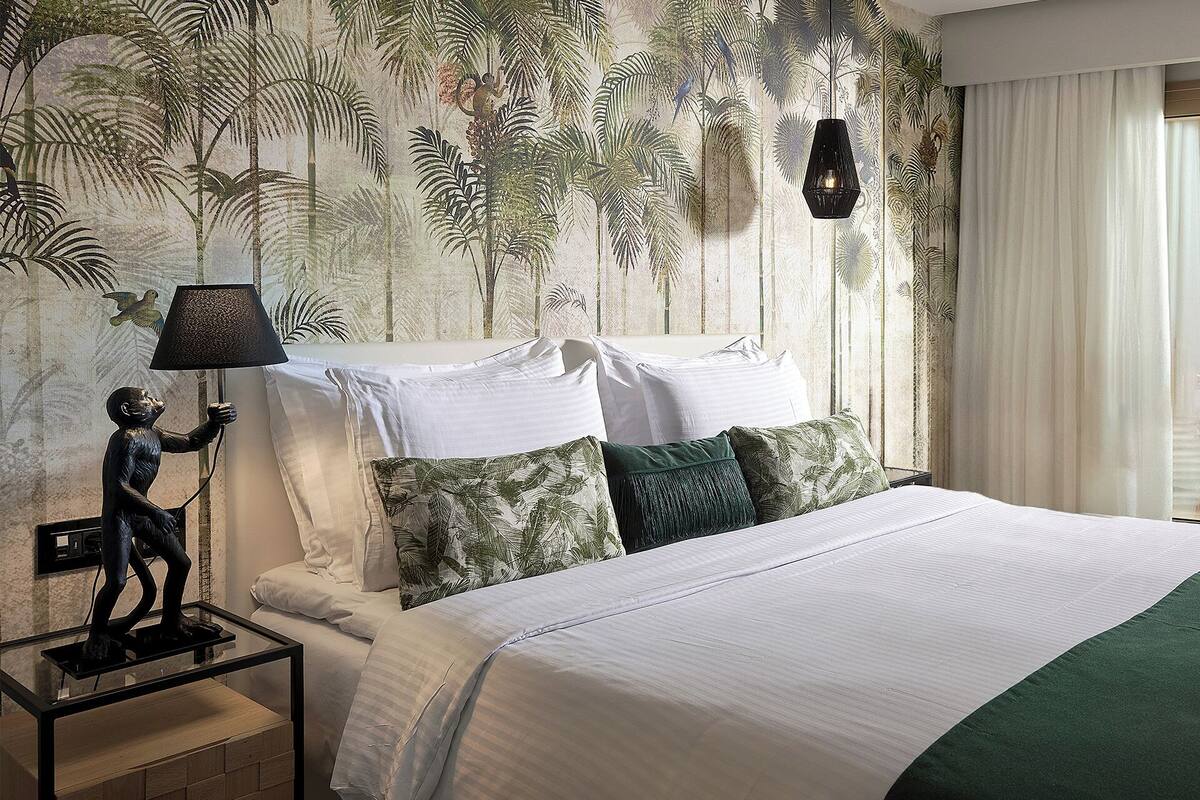 A bedroom with a bed, pillows, a nightstand with a lamp, and a wall with a tropical wallpaper.