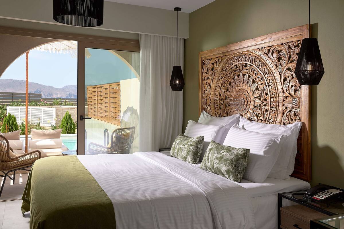 A hotel room with a large bed, a wooden headboard, a view of a pool, and a balcony with outdoor furniture.