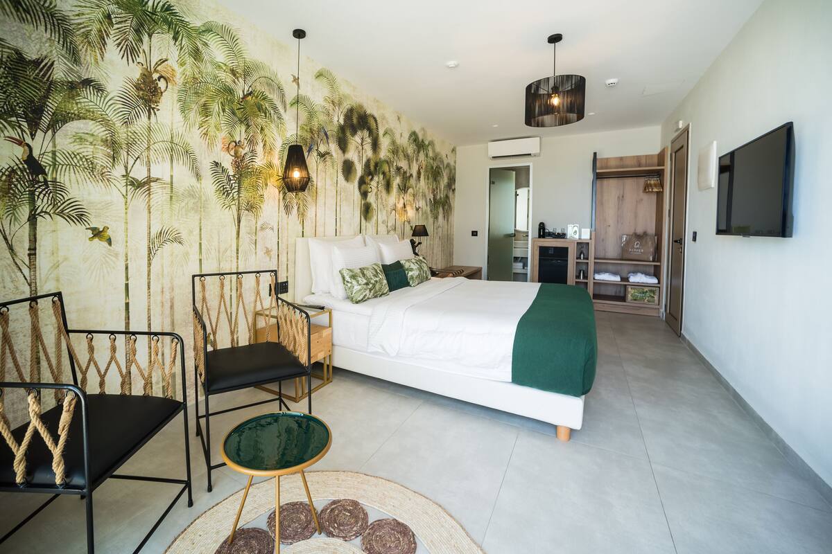 A bedroom with a bed, two chairs, a small table, a wall mural, a TV, and a wooden closet.