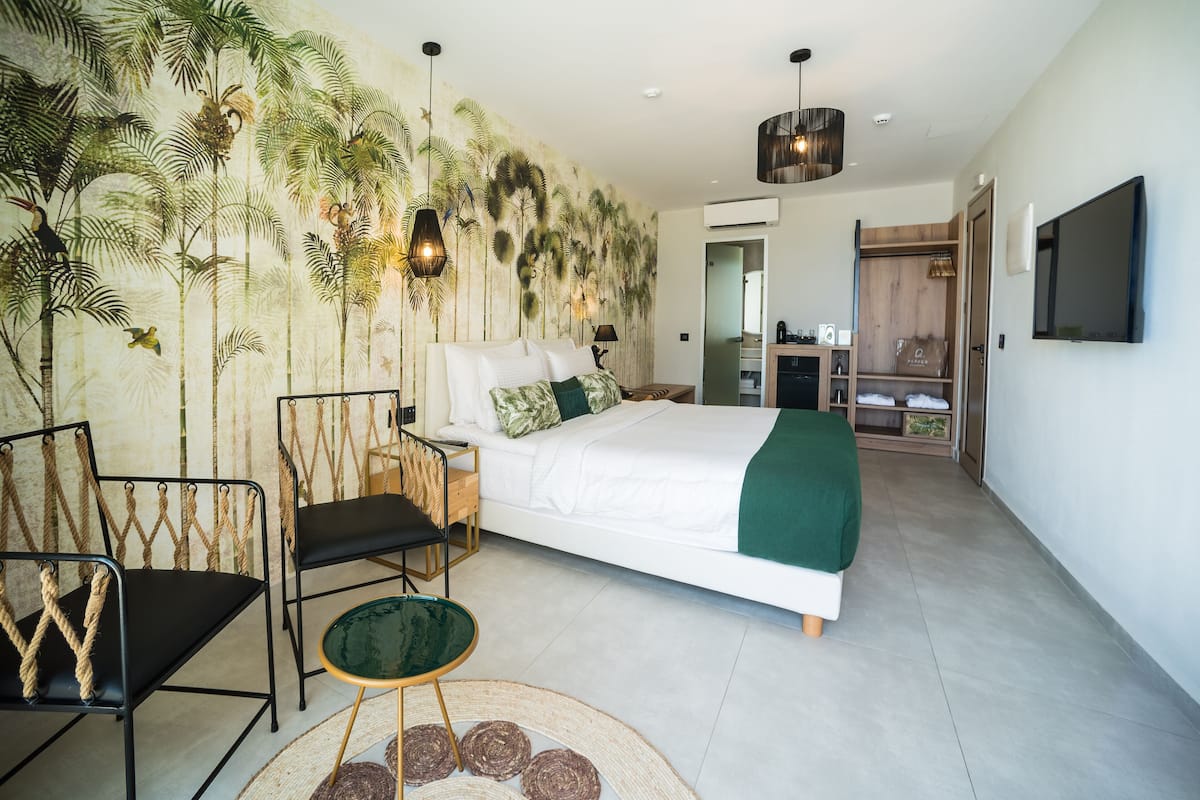 Quarto com cama, duas cadeiras, uma mesinha, um mural na parede, televisão e um guarda-roupa de madeira.