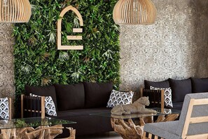 Una sala moderna con un sofá negro, cojines estampados, una mesa de centro de vidrio y una pared decorativa con una gran letra 'L'.