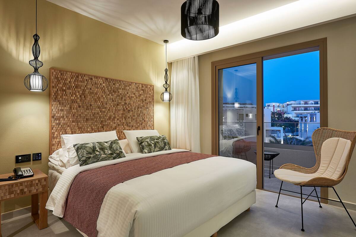 A modern hotel room with a large bed, a wooden headboard, a wicker chair, and a view of the city through a sliding door.