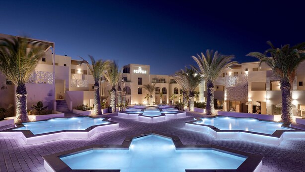 A modern hotel courtyard with multiple swimming pools, palm trees, and illuminated architecture at night.