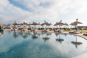 Ein Poolbereich mit Liegestühlen und Reetdachschirmen, ein klarer Himmel und ein ruhiges Meer im Hintergrund.