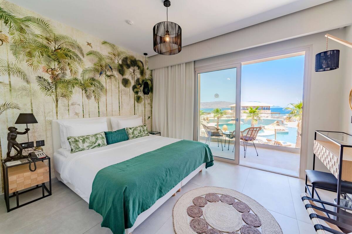 A bedroom with a bed, a nightstand, a lamp, a large window with a view of a pool and palm trees, and a ceiling light.