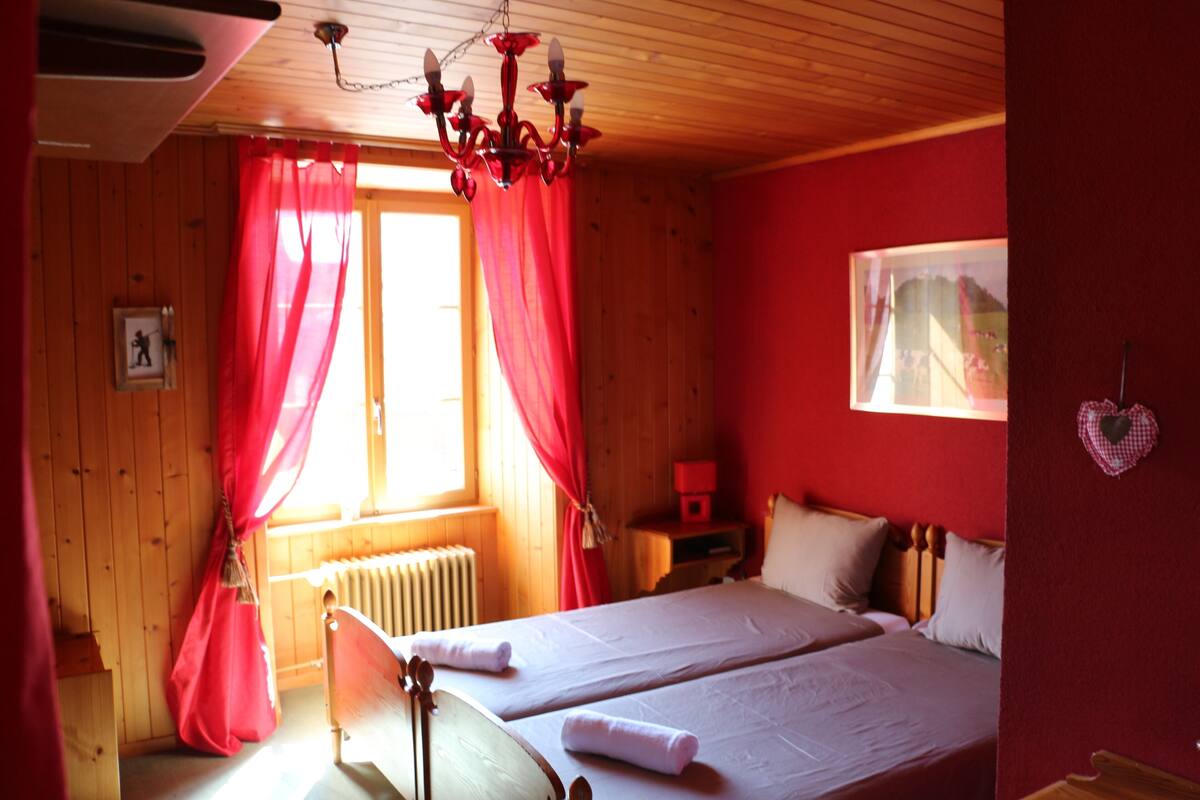 A room with wooden walls and a red accent wall, featuring two beds, a red chandelier, and a window with red curtains.