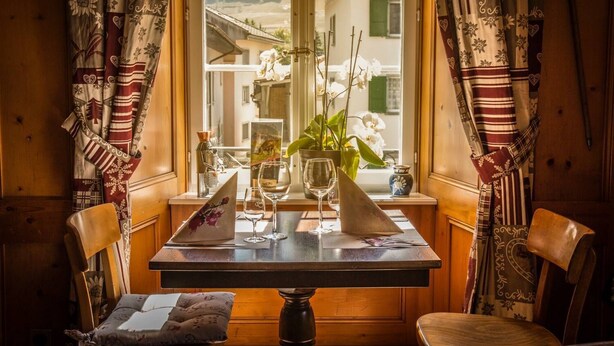 A cozy room with a wooden table set for two, a window with curtains, and a view of a building outside. - Auberge de l'Ange (Haut-Intyamon)