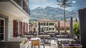 A terrace with outdoor seating, a view of a mountain, and traditional buildings in the background. - Auberge de l'Ange (Haut-Intyamon)