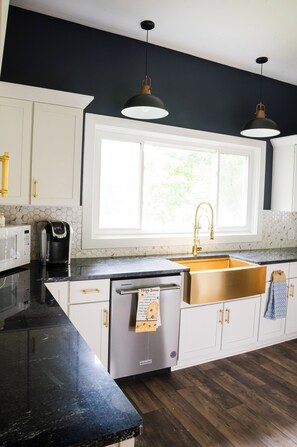 Una cocina moderna con armarios blancos, encimera negra, lavavajillas de acero inoxidable e iluminación colgante.