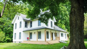 A two-story white house with a porch, surrounded by lush green trees. - Fall color tours!-Awesome Location! Luxury Farmhouse near Wineries & Orchards!! (Fennville)