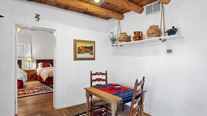 A traditional room with a wooden table, two chairs, a red and blue patterned rug, a painting on the wall, and a shelf with pottery and plants. - Casa Martinez | Historic Santa Fe Charm (Santa Fe)